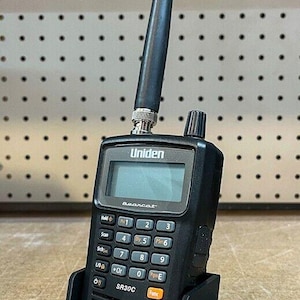 May include: A black Uniden Bearcat SR30C scanner radio with a black antenna and a black charging base. The radio has a digital display and a keypad with buttons labeled "Hold", "Scan", "S/S", "Pwr 1", "1", "2", "3", "4", "5", "Psrc 6", "7", "8", "9", "LO D", "Clr", "0", "Pgm E", "Func".