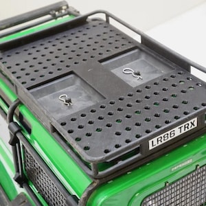May include: Black roof rack with a perforated design mounted on a green toy vehicle. The rack has a rectangular shape with raised edges and two recessed compartments. The vehicle has the text "LR86 TRX".