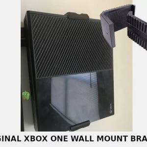 May include: A black Xbox One console mounted on a wall with two black wall mount brackets. The brackets are made of plastic and have a textured surface.