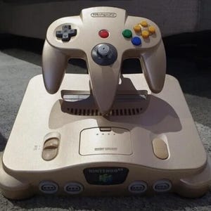 May include: A gold Nintendo 64 console with a matching gold controller. The console has a slot for game cartridges and two controller ports. The controller has a  "Nintendo" logo and colored buttons.