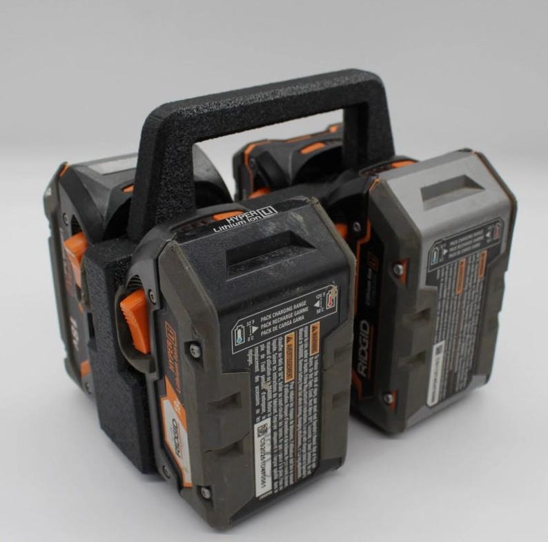 May include: A black and orange power tool battery holder with a textured handle. The holder contains multiple black and gray 18V batteries with orange accents. The batteries have printed labels and are designed for power tools.