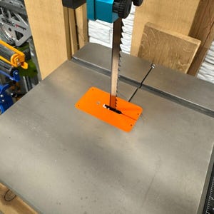 May include: A bandsaw with a silver blade and an orange safety guard. The saw has a blue and red motor housing and sits on a gray metal table. The background includes wooden panels and other workshop equipment.