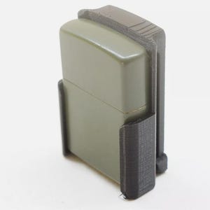 May include: A black plastic case designed to hold a lighter. The case has a textured surface and a green lighter is inside.