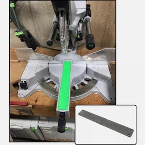 May include: A gray and green miter saw with a green handle and a gray base. The saw is on a wooden table. A gray rectangular plate is also shown.