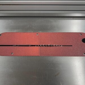 May include: A red table saw insert with a black oval hole and a long, thin slot for the saw blade. The insert is made of a durable material and is designed to fit a table saw.