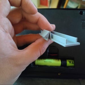 May include: A white plastic part with a triangular shape and two slots on the side. The part is being held by a hand.