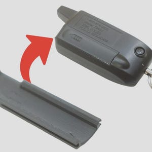 May include: A black car key remote with a detached gray plastic cover. The remote has a key ring and a button. The cover is rectangular with a curved edge. The image shows the remote and cover separated by a red arrow.