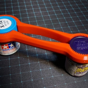 May include: Orange plastic tool for opening paint bottles. The tool has two circular openings, each with a different size and shape. The tool is holding two paint bottles, one blue and one purple.