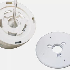 May include: White plastic security camera housing with a round base and a separate white plastic mounting plate. The camera housing has a circular opening in the center and a series of small holes around the edge. The mounting plate has three small holes around the edge and a larger circular opening in the center.