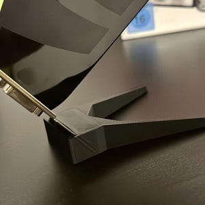May include: A black plastic stand with a triangular shape, designed to hold a phone or tablet upright. The stand is made of a durable material and has a sleek, modern design.