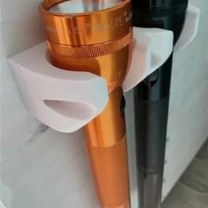 May include: Two flashlights mounted on a white wall mount. The flashlight on the left is orange and says "MAG-LITE" on the side. The flashlight on the right is black and says "MAG-LITE" on the side.