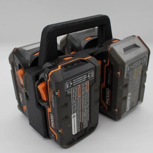 May include: A black battery holder with a black handle, holding four black and orange Ridgid 18V Lithium-Ion batteries. The batteries have orange release buttons and white text. The holder is designed to carry multiple batteries.