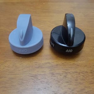 May include: Two stove knobs, one light blue and one black, sit on a wooden surface. The light blue knob has the word "OFF" printed on it. The black knob has the word "OFF" and "LITE" printed on it.