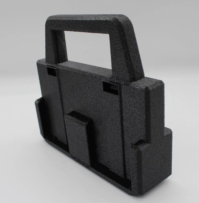 May include: A textured black plastic carrying case with a handle. The case has a rectangular shape with a handle on top and a series of slots and protrusions on the sides. The surface has a slightly rough, matte finish.