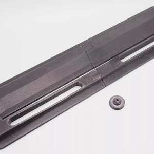 May include: A black plastic part with a long, narrow rectangular shape and a small, round, black plastic piece. The part has several grooves and holes.