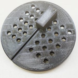 May include: A black 3D printed game board with a grid of holes and a black rectangular piece on top. The board is divided in half by a thin line.