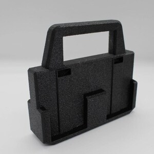 May include: A textured black rectangular object with a handle. The object has a boxy shape with several protruding rectangular features. The handle is a rectangular opening at the top of the object.