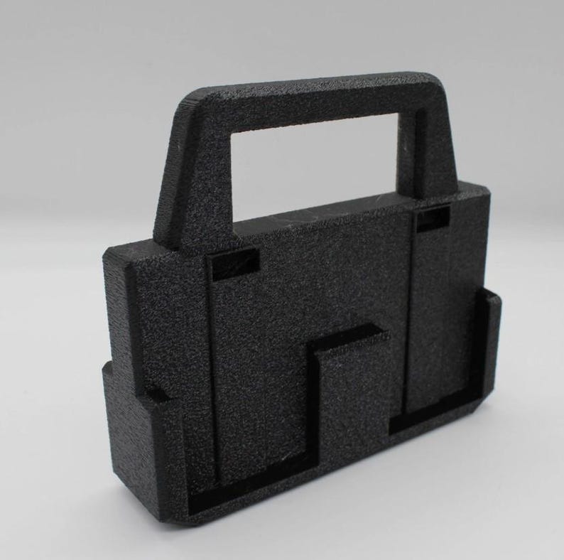 May include: A textured black rectangular object with a handle. The object has a boxy shape with several protruding rectangular features. The handle is a rectangular opening at the top of the object.