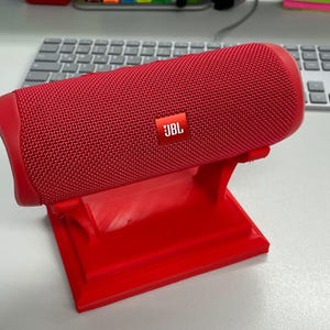 May include: A red JBL portable Bluetooth speaker on a red 3D printed stand. The speaker has a textured surface and the JBL logo is visible on the front.