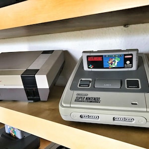May include: A gray Nintendo Entertainment System and a gray Super Nintendo Entertainment System with a Super Mario World game cartridge inserted. Both consoles are on a wooden shelf.