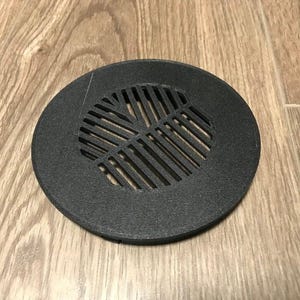 May include: A round, black drain cover with a slotted design. The cover is made of a matte material and sits on a wooden surface. The slotted pattern allows for water drainage.