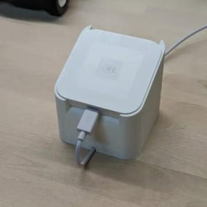 May include: A white, square-shaped device with a USB cable attached. The device has a flat top with a subtle logo and rounded corners. The cable is plugged into the front of the device.
