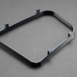 May include: A dark gray, rectangular frame with rounded corners and small protrusions on the sides. The frame is made of a solid material and sits on a gray surface. The object appears to be a component or part.
