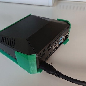 May include: A black computer with a green protective case. The computer has multiple ports on the back, including USB and ethernet. A black cable is connected to the computer.