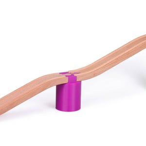 May include: A wooden ramp with a curved design, supported by two colorful cylindrical blocks. The blocks are stacked, with a purple block on top of a green block. The ramp is made of natural wood and has a smooth surface.