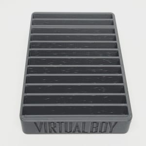 May include: A dark gray plastic organizer with multiple horizontal slots. The base of the organizer has the text "VIRTUAL BOY" embossed on it. The object is designed to hold items and is set against a white background.