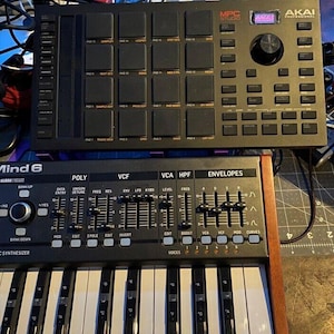 May include: A black and silver Akai MPC Studio music production controller with 16 pads and a large knob. A black and silver Mind 6 synthesizer with a 25-key keyboard and knobs for poly, VCF, VCA, HPF, and envelopes.