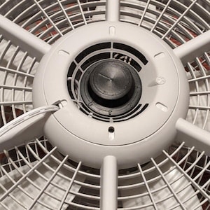 May include: Close-up of a white oscillating fan. The fan has a circular design with a central control knob and a protective grill. The fan blades are visible behind the grill.