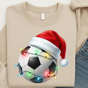 Könnte beinhalten: Beiges Sweatshirt mit einem Fußball, der einen roten Weihnachtsmannhut mit weißem Rand trägt und mit bunten Lichterketten umwickelt ist. Der Fußball hat schwarz-weiße Paneele.