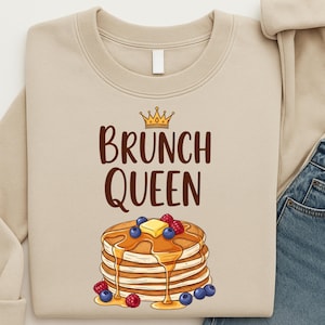 Könnte beinhalten: Beiges Sweatshirt mit dem Text "BRUNCH QUEEN" in Braun, gekrönt von einer goldenen Krone. Darunter ein Stapel Pfannkuchen mit Sirup, Butter, Blaubeeren und Himbeeren.