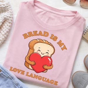 Könnte beinhalten: Hellrosa T-Shirt mit dem Text "BREAD IS MY LOVE LANGUAGE". Das Design zeigt eine Comic-Zeichnung eines Toastbrots, das ein rotes Herz umarmt. Das Brot hat ein lächelndes Gesicht.