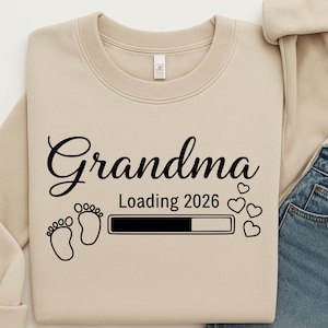May include: A beige sweatshirt with the word "Grandma" in black script. Below, the text "Loading 2026" appears with a loading bar, footprints, and heart graphics. The sweatshirt is folded with a portion of blue jeans visible.