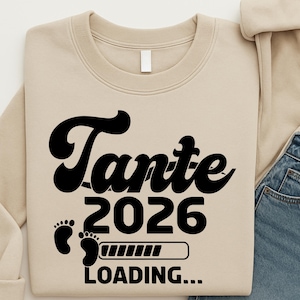 May include: A beige sweatshirt with the word "Tante" in a black, stylized font. Below, the text "2026" is displayed, along with baby footprints and a loading bar graphic. The word "LOADING..." is at the bottom.