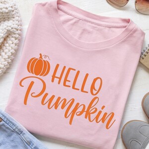 Könnte beinhalten: Hellrosa T-Shirt mit dem orangefarbenen Schriftzug "HELLO Pumpkin" und einer Kürbisgrafik. Das Shirt hat einen Rundhalsausschnitt und ist aus weichem Material. Das Design befindet sich mittig auf der Vorderseite.