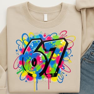Pu&ograve; includere: Felpa beige con un vivace design in stile graffiti del numero 67. Il numero &egrave; delineato in nero e riempito con uno spruzzo colorato di vernice blu, verde, gialla e rosa. Il design ha un'estetica street art.