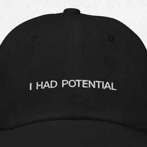 Pode incluir: Boné de beisebol preto com a frase bordada em branco "I HAD POTENTIAL". O boné tem uma aba curva e uma coroa arredondada. O texto está centralizado.