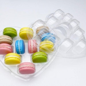May include: A heart-shaped clear plastic tray containing an assortment of colorful macarons in shades of green, yellow, pink, blue, and tan. The tray features individual molded compartments for each macaron, with a hinged lid partially open.