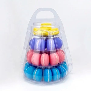 May include: A clear plastic tiered display stand containing four levels of round macarons. The top tier holds yellow macarons, the second purple, the third pink, and the bottom tier blue macarons. The stand features a handle at the top.
