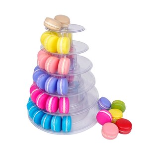 May include: A multi-tiered clear plastic display stand arranged in a conical shape, holding rows of colorful macarons in shades of beige, yellow, pink, purple, magenta, and blue, with additional loose macarons placed at the base of the stand.