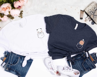 Può includere: Due magliette, una bianca e una blu navy, ciascuna con un design a tasca con un animale dei cartoni animati. La maglietta bianca ha un panda e la maglietta blu navy ha un orso. Entrambe le magliette sono abbinate a jeans in denim e scarpe da ginnastica bianche.