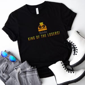 May include: Black t-shirt with a yellow and orange graphic of a robot wearing a crown. The text "Nicholas King of the Losers!" is printed below the graphic.