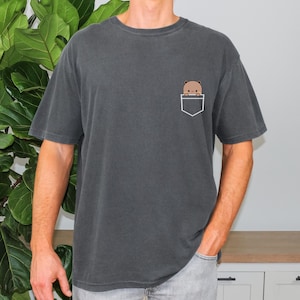 May include: A dark grey t-shirt with a small pocket on the left side. A cartoon bear is peeking out of the pocket.