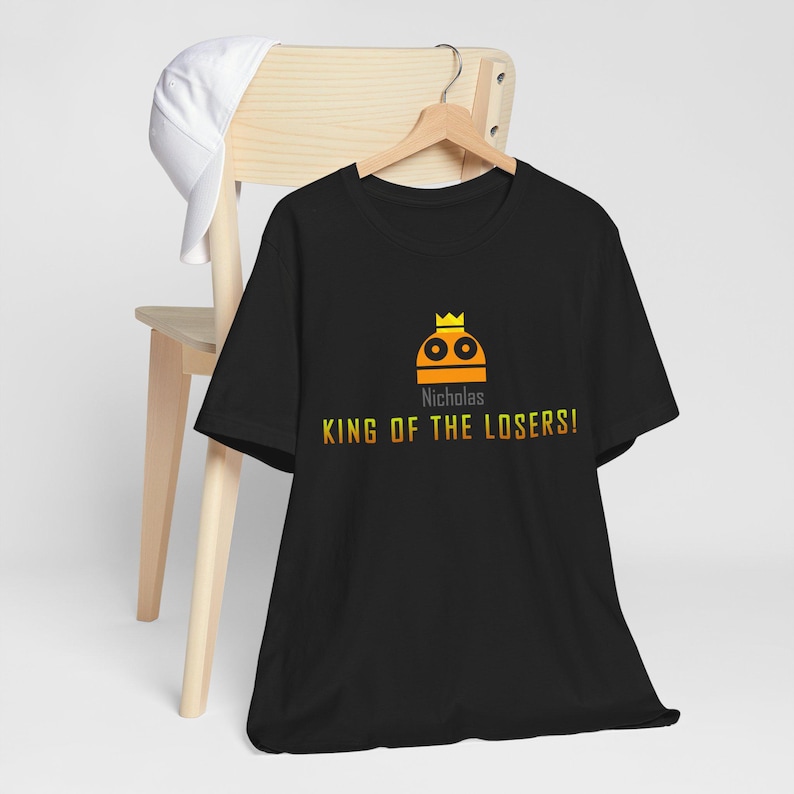 May include: Black t-shirt with a yellow and orange graphic of a robot wearing a crown. The text "Nicholas" and "KING OF THE LOSERS!" is printed on the shirt.