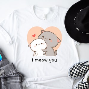 K&ouml;nnte beinhalten: Wei&szlig;es T-Shirt mit einem Cartoon-Bild von zwei Katzen, die sich in einem Herzen umarmen. Der Text "i meow you" ist unter dem Bild gedruckt.