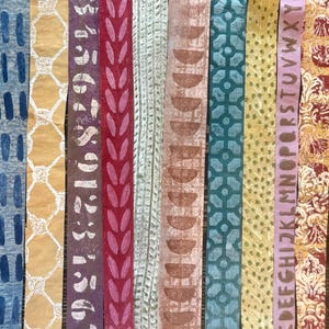 May include: A collection of ten vertical paper strips with various patterns and colors. Designs include blue dashes, yellow lattice, purple numbers, red leaves, and brown half-circles. Other strips feature green geometric shapes, yellow dots, and a lavender strip with the alphabet.