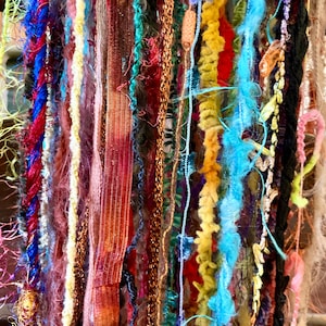 May include: A collection of multi-textured yarn and ribbon strands in a vibrant array of colors including bright blue, deep red, mustard yellow, copper, and magenta. The assortment features various thicknesses, fuzzy textures, and metallic finishes.